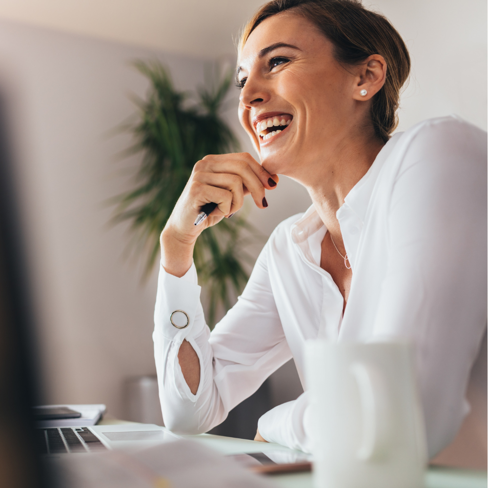 Smiling woman in a white blouse, resting her chin on her hand while looking thoughtfully engaged at a desk with a laptop and a coffee mug.