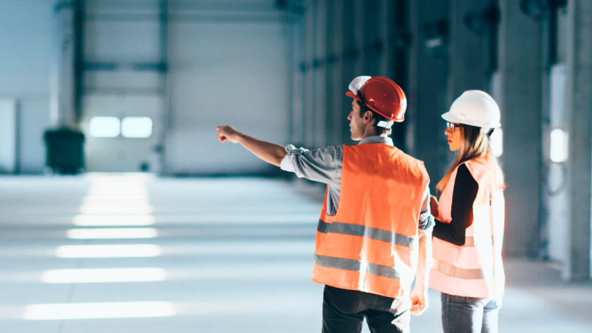 Two construction workers, one in a red hard hat and the other in a white hard hat, discuss plans inside a large, empty warehouse. They are wearing high-visibility vests and standing in a sunlit area with shadows cast on the floor.